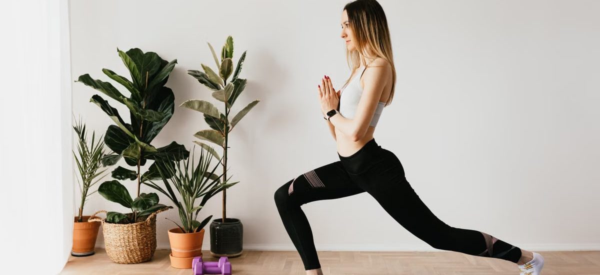 Side view of fit lady in sports clothes and wristwatch standing in Anjaneyasana pose while practicing yoga on mat near dumbbells and potted plants at home and looking away